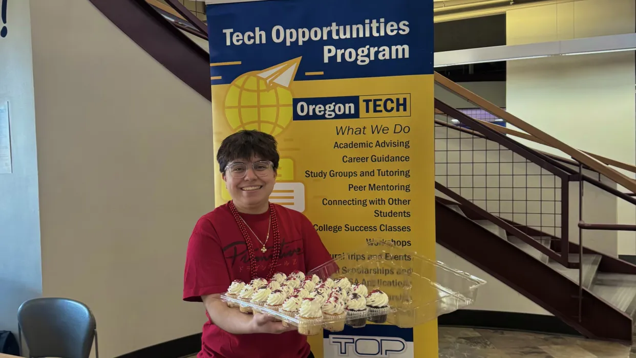 Student standing in front of TOP informational banner holding a tray of cupcakes