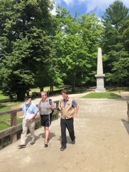 Audio Description researcher Brett Oppegaard, on the viewer's right, uses an audio recorder to capture the thoughts of Bob Hachey, center, as they walk across the North Bridge with NPS staff member Steve Neth at Minute Man National Historical Park near Concord, MA.